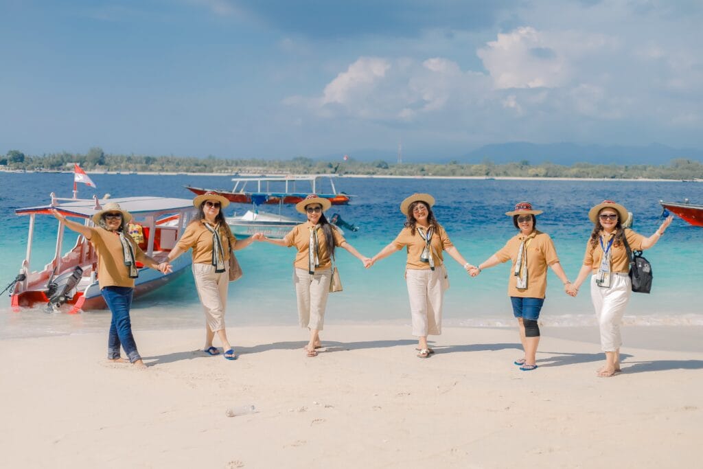 Enam orang wanita berseragam outing perusahaan sedang bergandengan tangan di tepi Pantai Gili Trawangan.