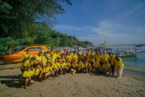 Sebuah rombongan besar wisatawan mengenakan seragam kuning berfoto bersama di tepi pantai dekat pelabuhan di Lombok.