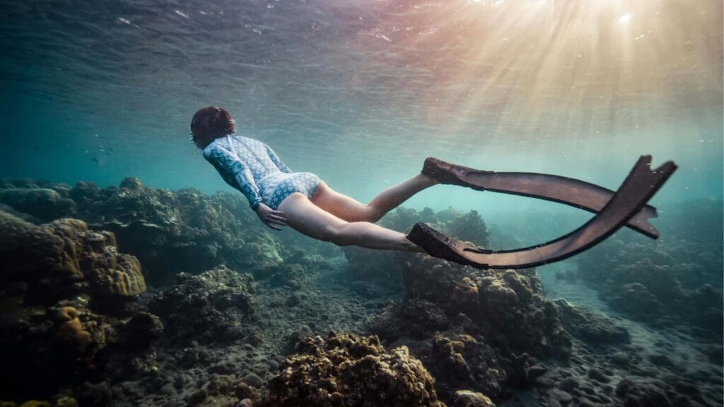 Seorang wanita sedang melakukan freediving di antara terumbu karang dengan sinar matahari yang menembus permukaan air di Gili, Lombok