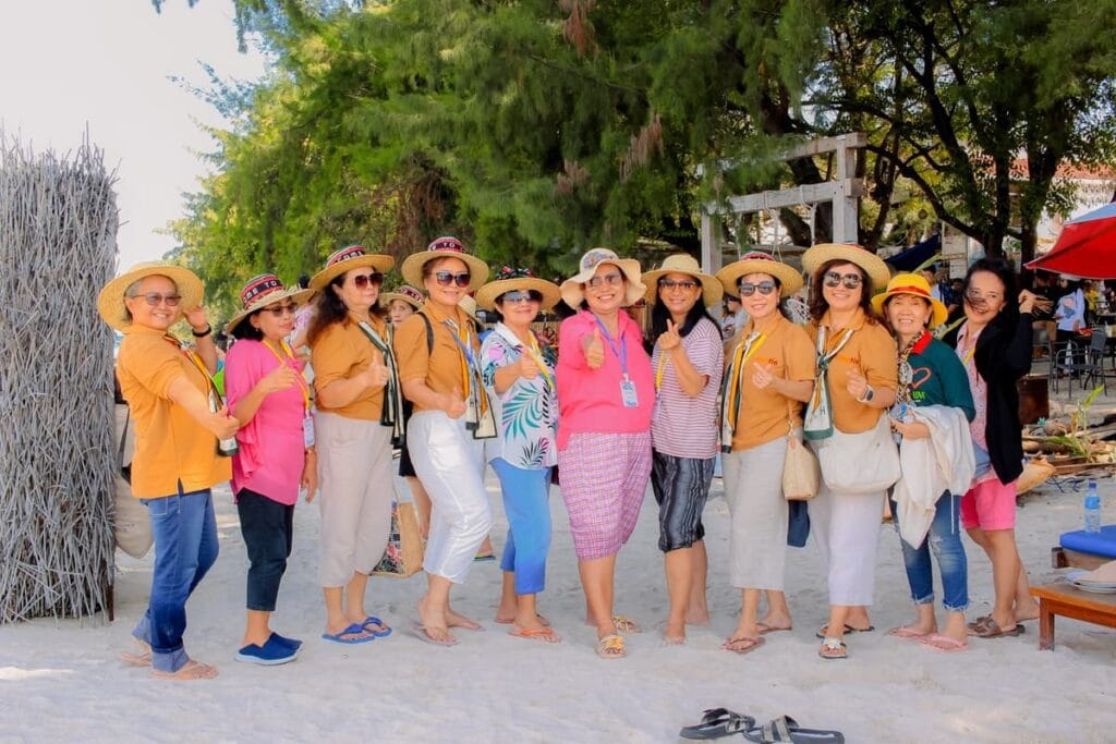 Sekelompok wisatawan mengenakan pakaian santai dan topi pantai berfoto bersama di area pantai berpasir putih dengan pepohonan rindang di belakangnya