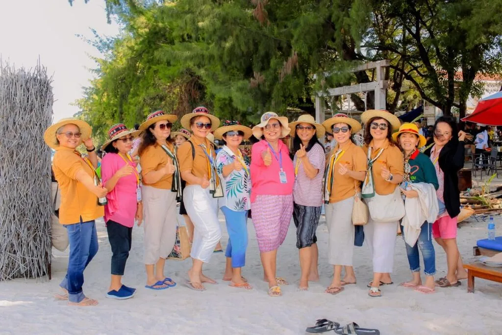 Sekelompok wisatawan mengenakan pakaian santai dan topi pantai berfoto bersama di area pantai berpasir putih dengan pepohonan rindang di belakangnya