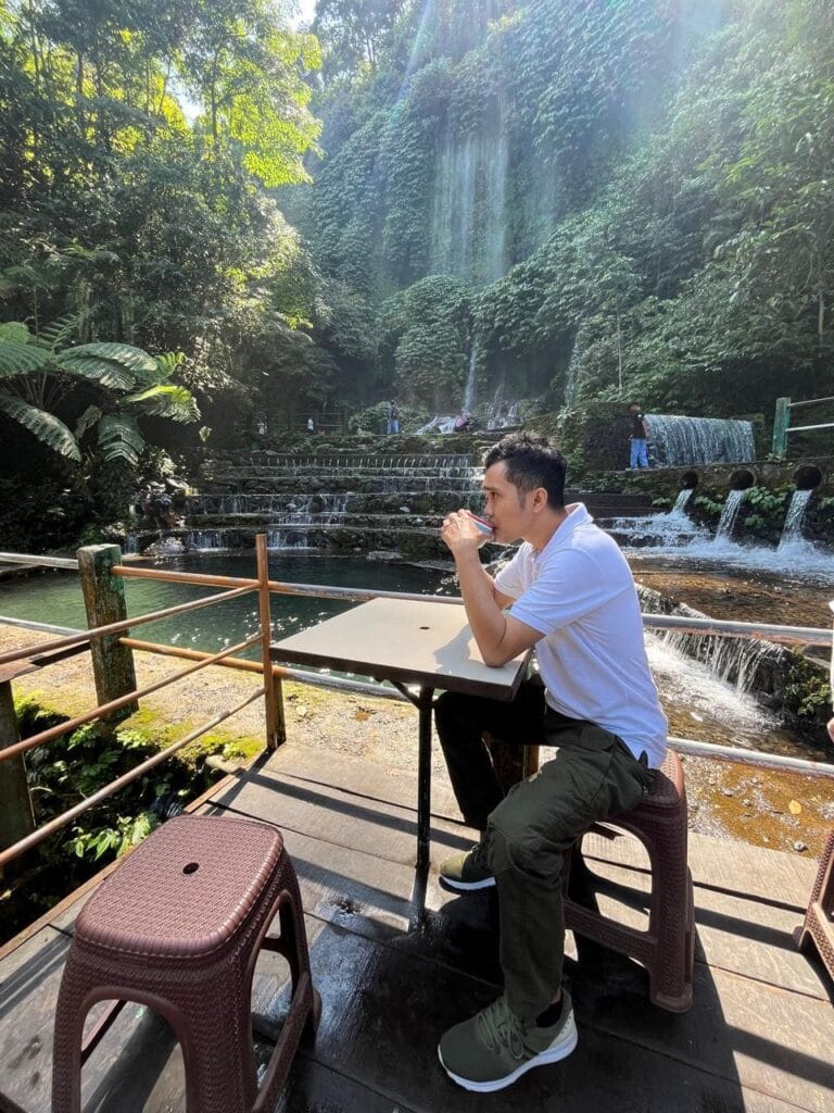 Seorang pria berbaju putih sedang duduk santai di cafe tepi air terjun Benang Kelambu, Lombok, menikmati pemandangan alam yang hijau dan asri.