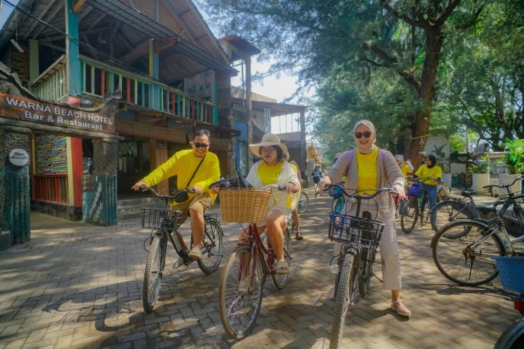 Tiga wisatawan sedang menikmati aktivitas bersepeda keliling pulau Gili Trawangan di jalanan yang rindang.