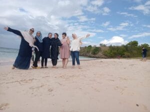 Sekelompok wisatawan berdiri di tepi Pantai Pink Lombok, berpose ceria dengan latar pasir berwarna cerah, langit biru, dan bukit hijau di kejauhan