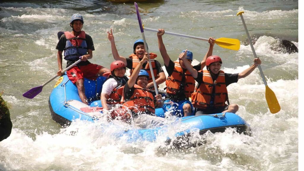 Sekelompok orang ceria melakukan arung jeram di atas perahu karet biru, mengarungi jeram sungai yang deras sambil mengangkat dayung mereka dengan gembira.