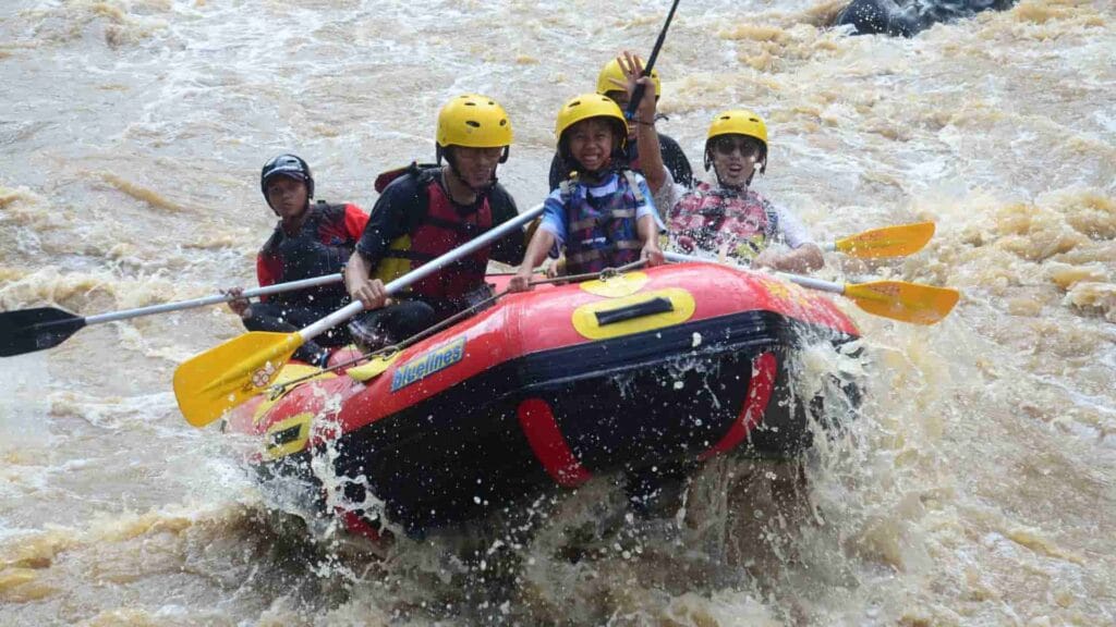 Sekelompok wisatawan yang ceria dengan helm kuning tersenyum lebar ke kamera dari atas perahu arung jeram merah, saat menerjang jeram di sungai yang airnya berwarna cokelat.