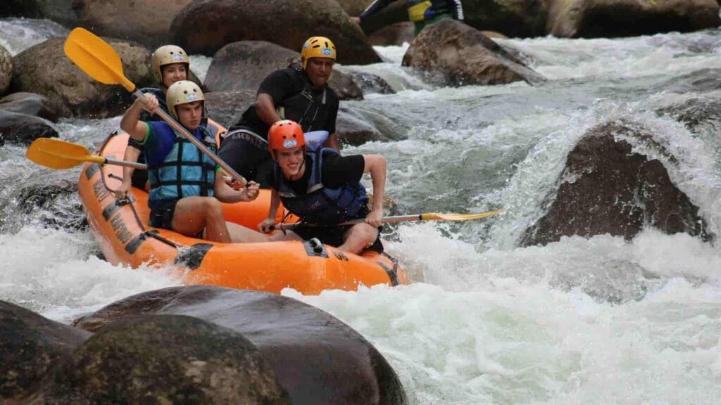 Tiga wisatawan dan satu pemandu di perahu arung jeram oranye sedang bermanuver secara teknis di antara jeram sungai yang deras dan berbatu-batu besar.